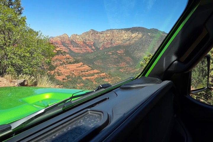 View from a green car's interior of red rock mountains and trees under a clear blue sky.