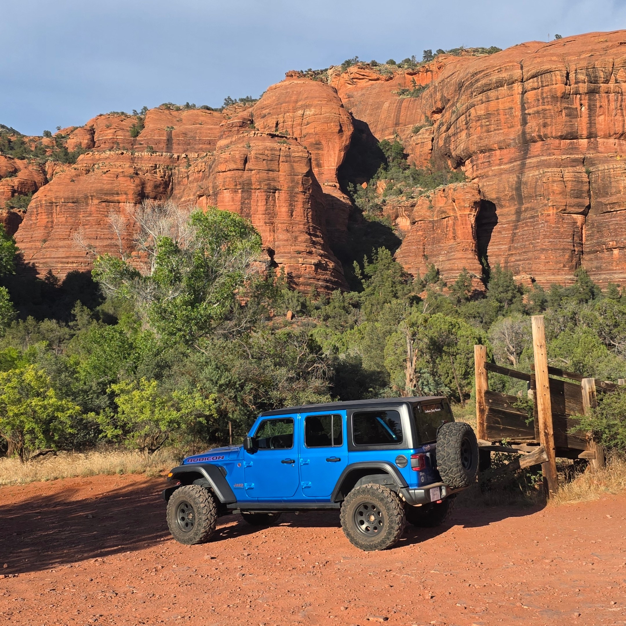 Blue jeep parked in front of red rock cliffs and greenery.