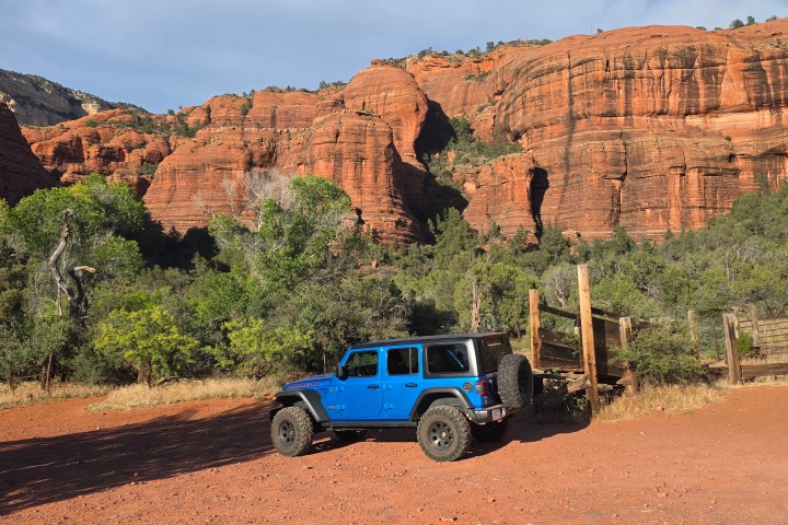 Blue jeep parked in front of red rock cliffs and greenery.