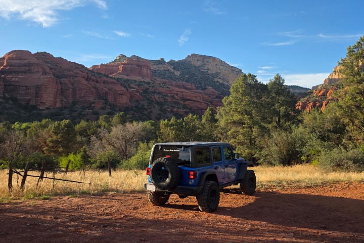 Blue SUV parked on dirt road in front of red rock formations and trees under a blue sky.