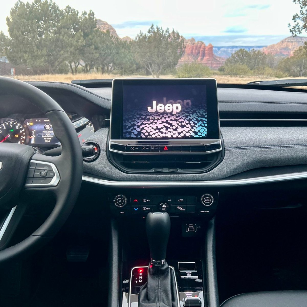 Jeep dashboard view with steering wheel, infotainment screen, and scenic background.
