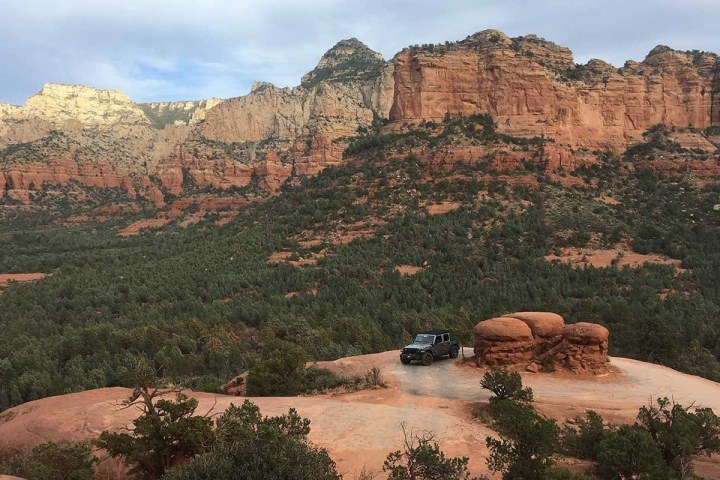 Jeep on rocky terrain with red cliff backdrop and green forest.